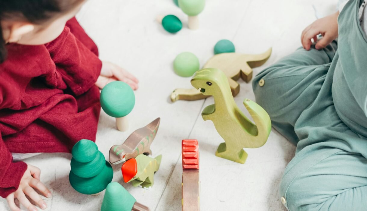 Two children engaged in imaginative play with wooden dinosaur toys on the playroom floor.