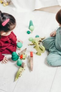 Two children engaged in imaginative play with wooden dinosaur toys on the playroom floor.
